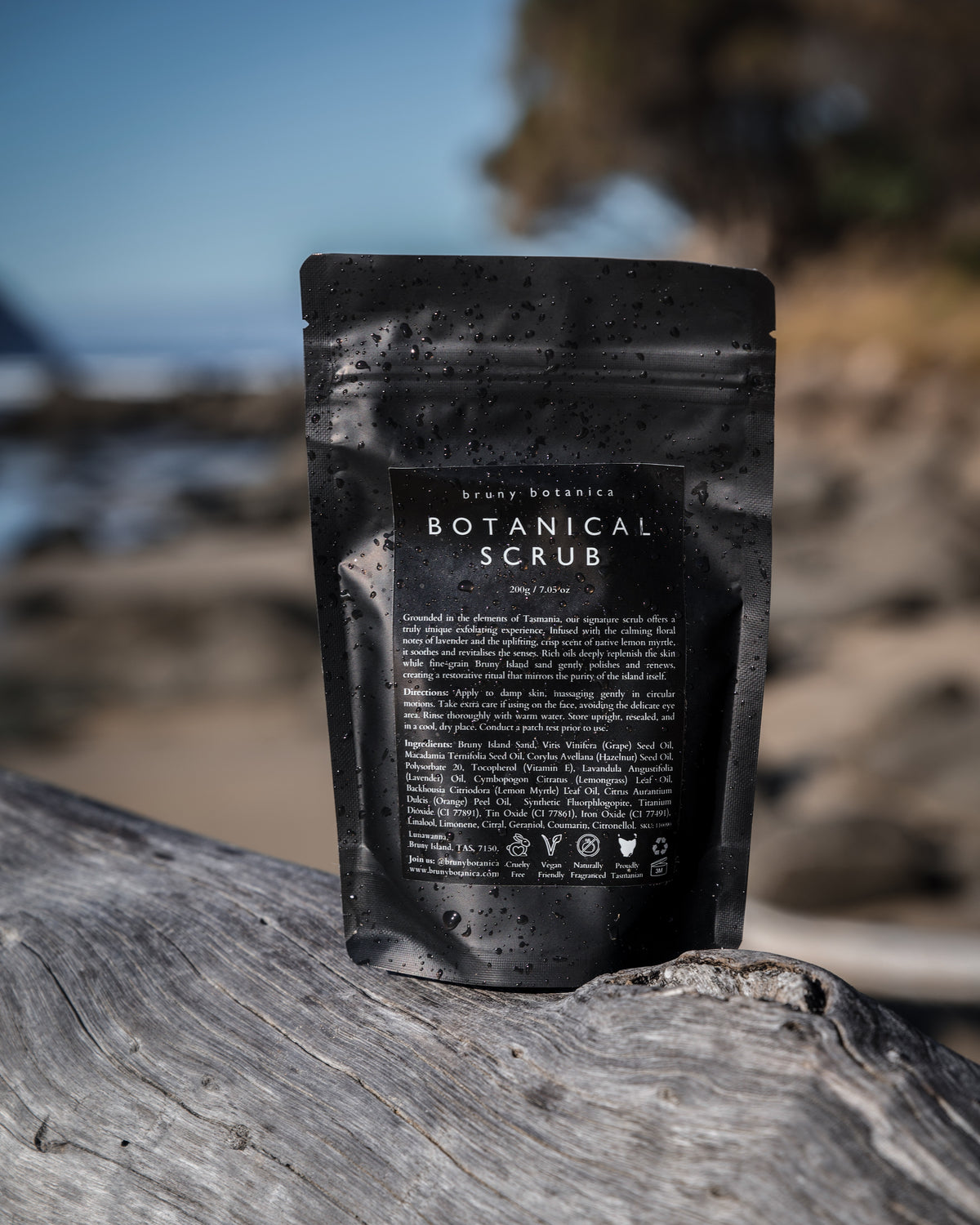 A black stand-up pouch of Bruny Botanica Botanical Scrub with water droplets, standing on a weathered log with a blurred beach background.