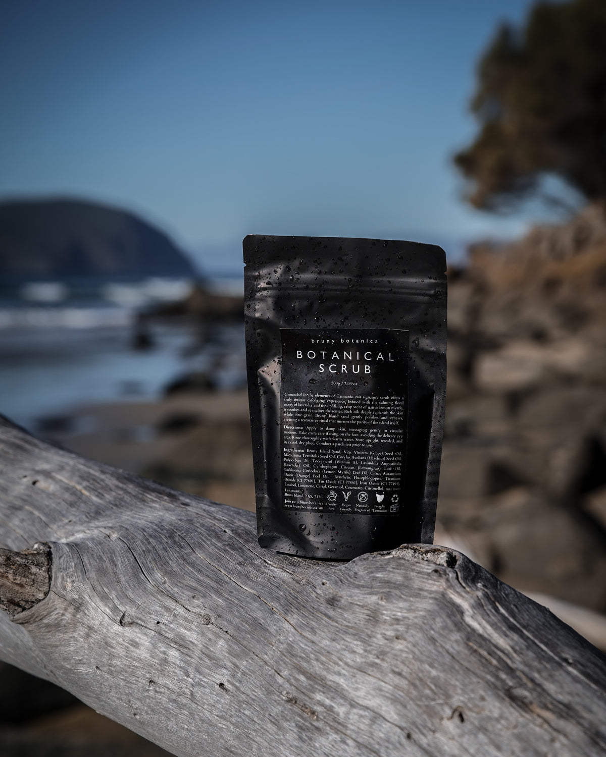 A black stand-up pouch of Bruny Botanica's Botanical Scrub standing on a weathered log with a blurred beach and rocky shoreline in the background.