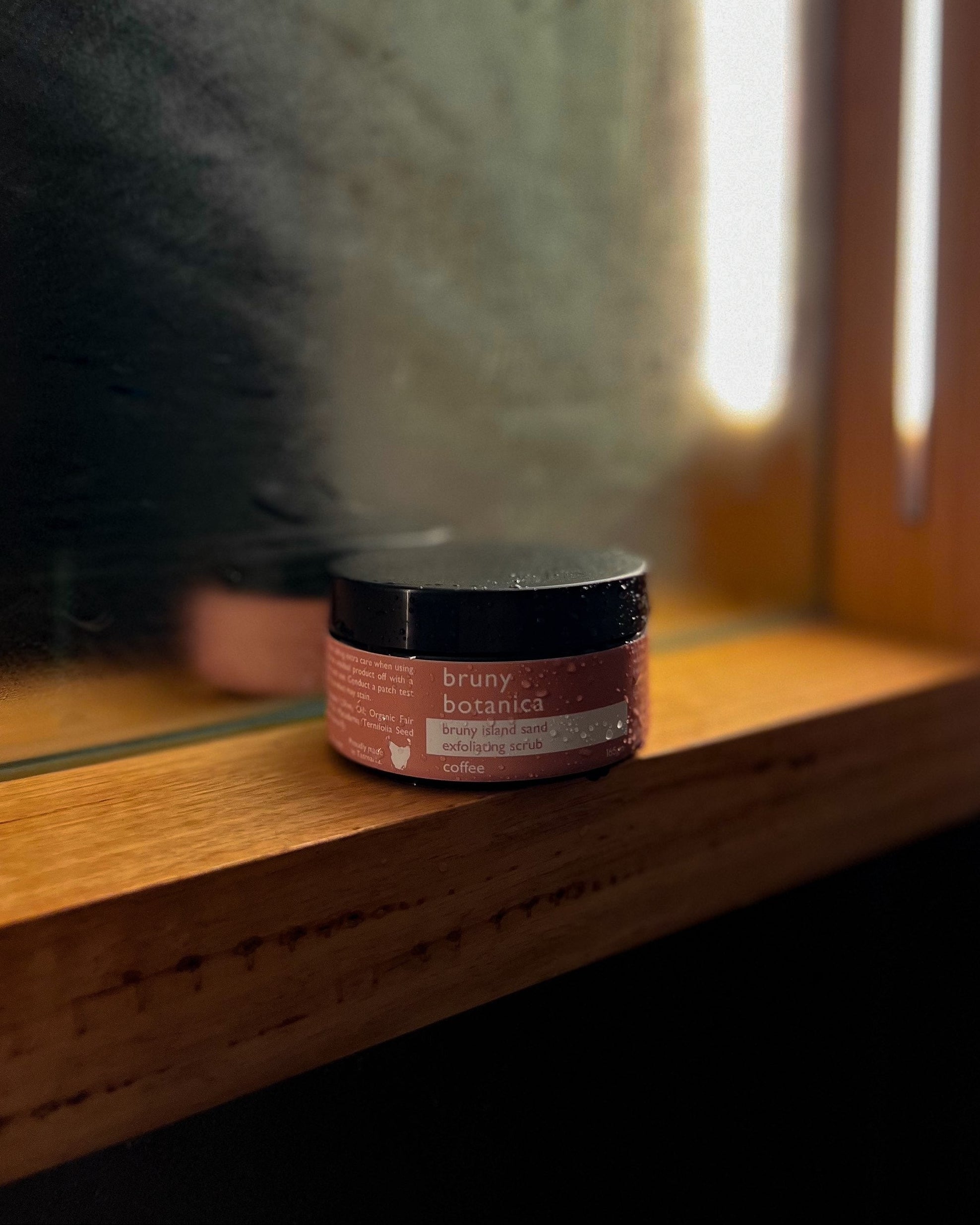 Small container of Bruny Island Sand Exfoliating Body Scrub - Coffee on a wooden ledge with a mirror in the background