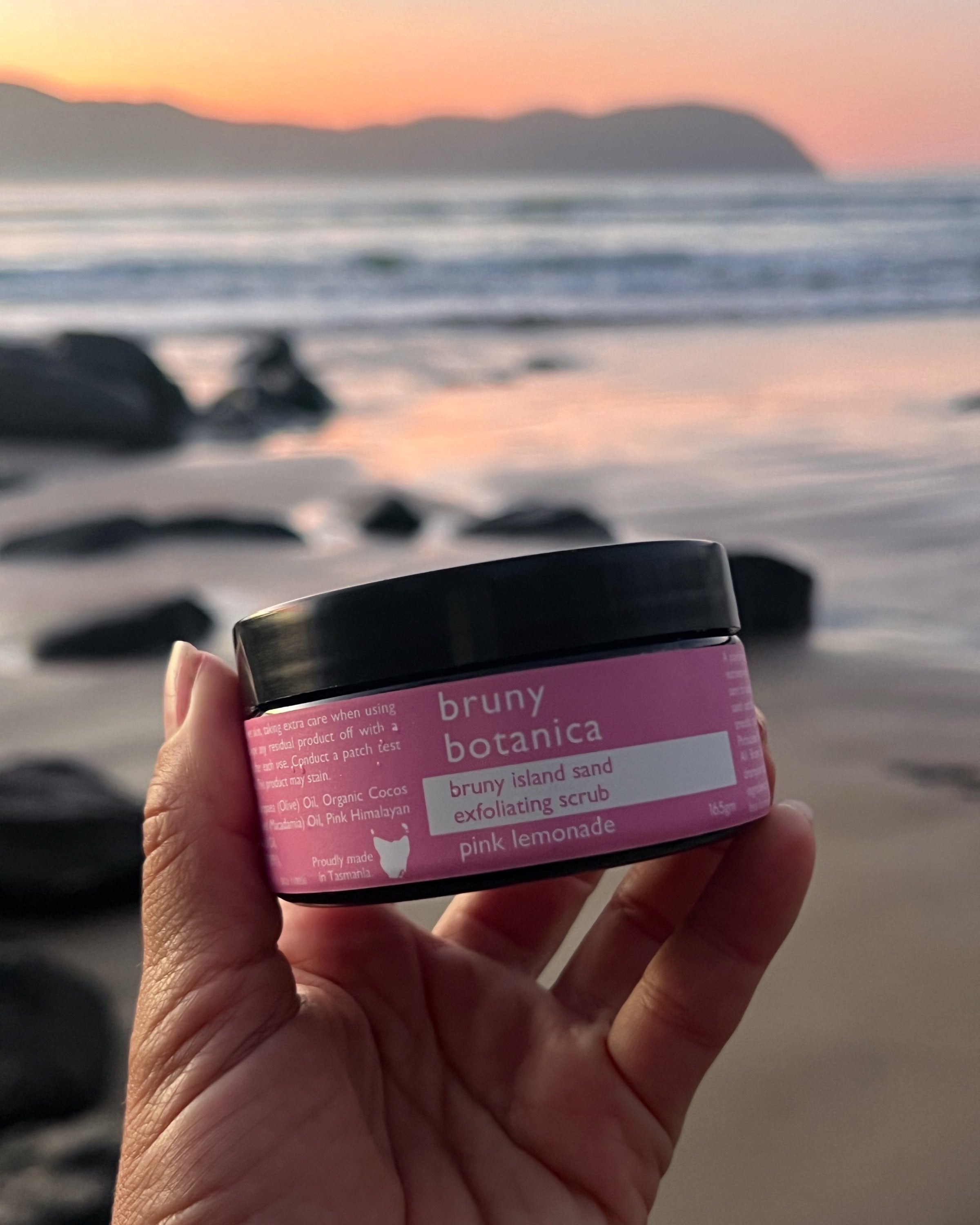 Hand holding a jar of Bruny Island Sand Exfoliating Body Scrub - Pink Lemonade against a beach sunrise at Cloudy Bay, Bruny Island