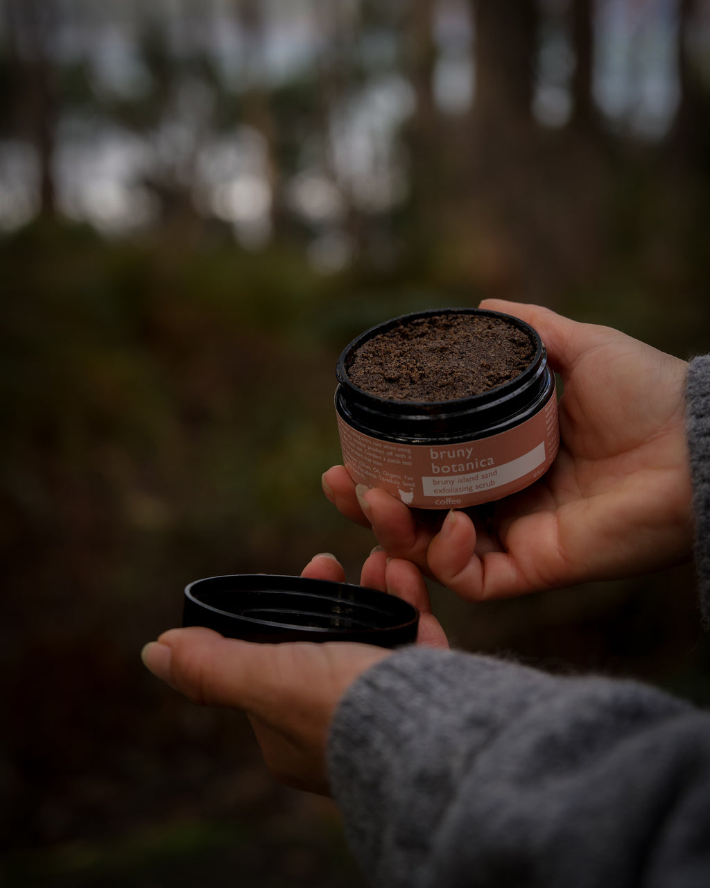 Hands hold open a jar of Bruny Island Sand Exfoliating Scrub Coffee against a forest background