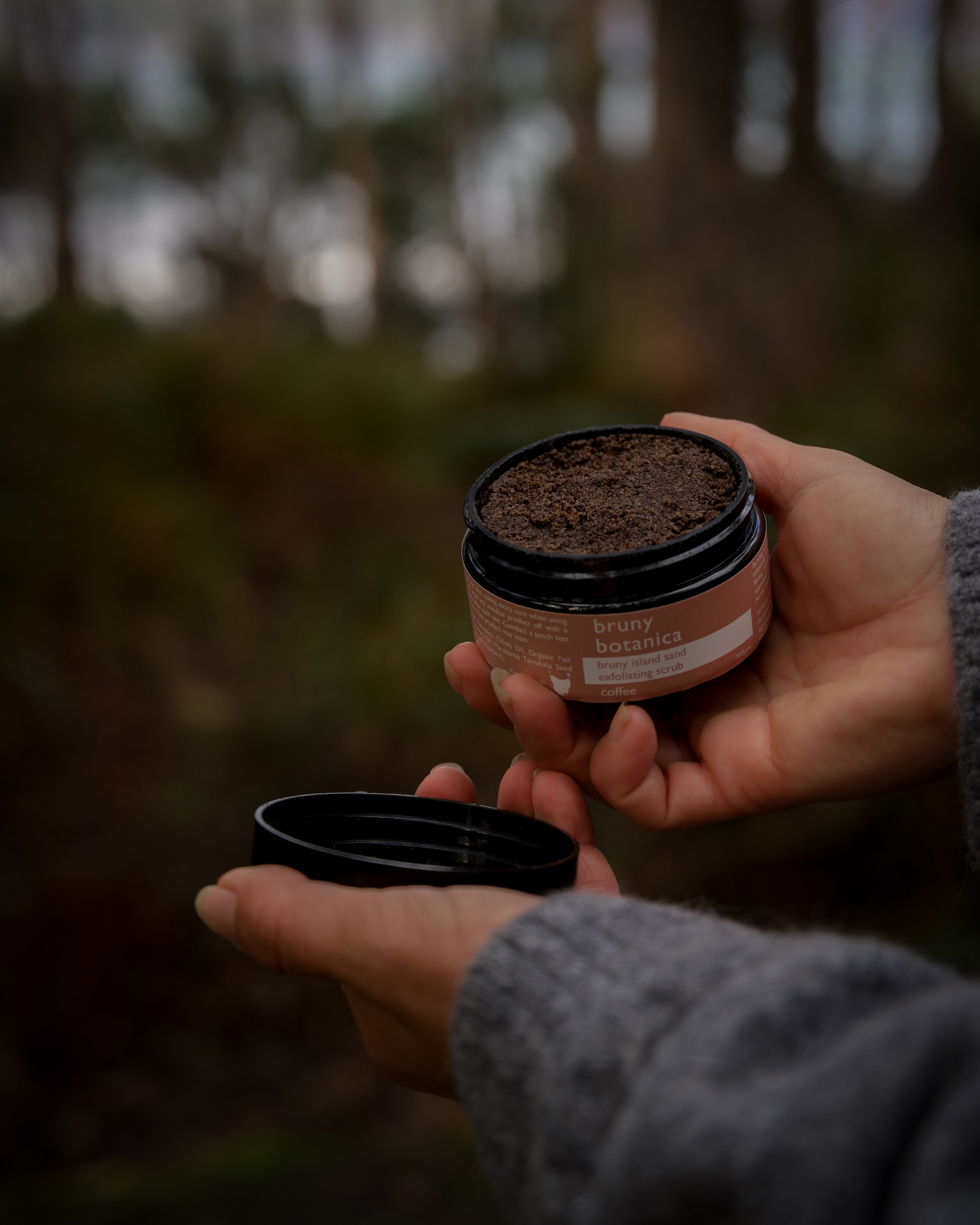 Hands hold open a jar of Bruny Island Sand Exfoliating Scrub Coffee against a forest background