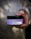 A hand holds a Bruny Island Sand Exfoliating Body Scrub - Lavender out towards the camera