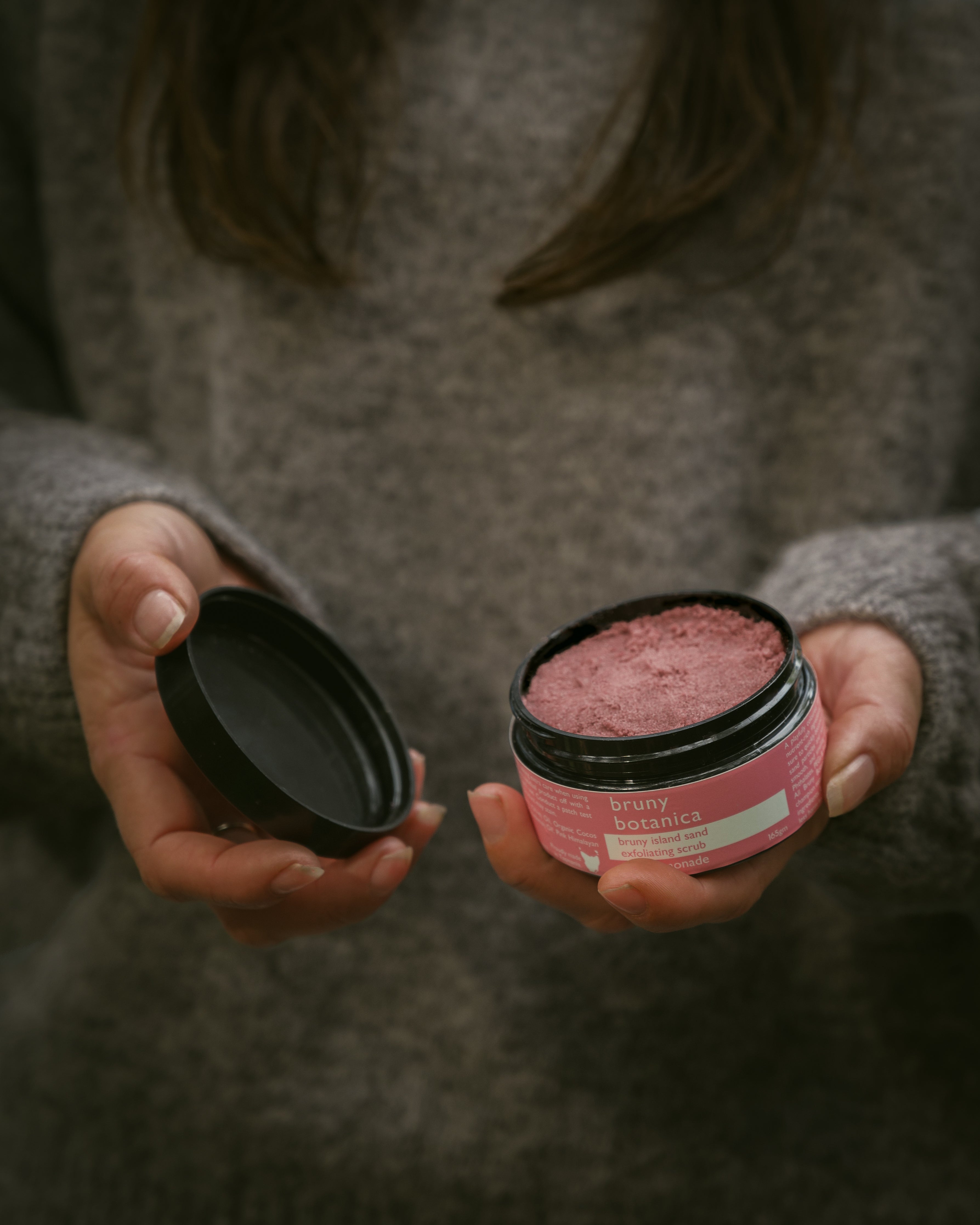 Two hands hold open a jar of Bruny Island Sand Exfoliating Scrub Pink Lemonade, showing the pink body scrub inside.