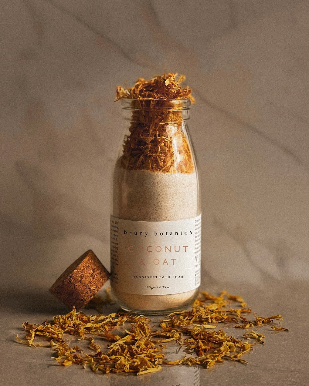 Bottle of Coconut & Oat Magnesium Bath Soak with a cork lid and scattered calendula flowers on a neutral background