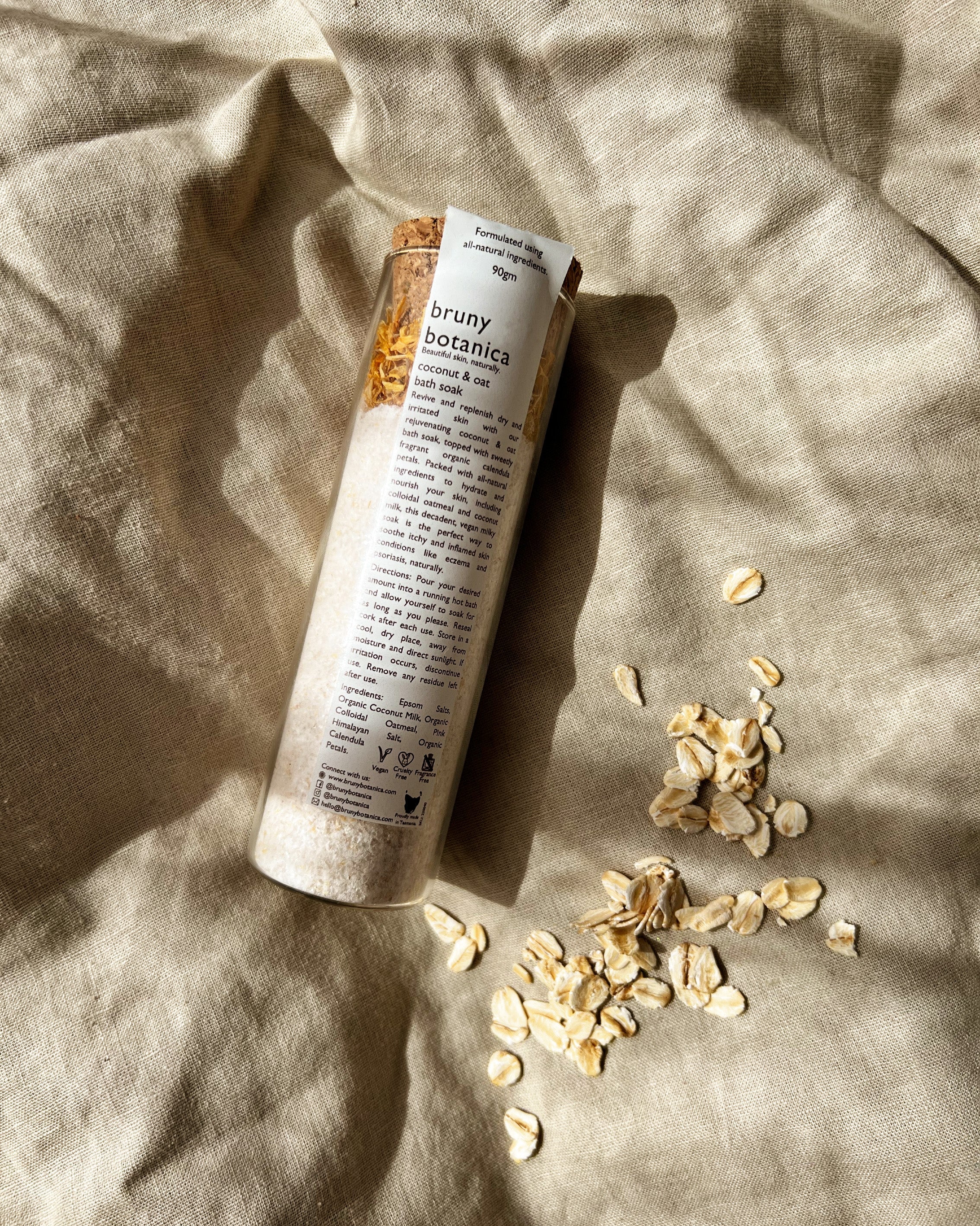 Bruny Botanics Coconut & Oat Magnesium Bath Soak tube on a textured beige fabric background with scattered oats.