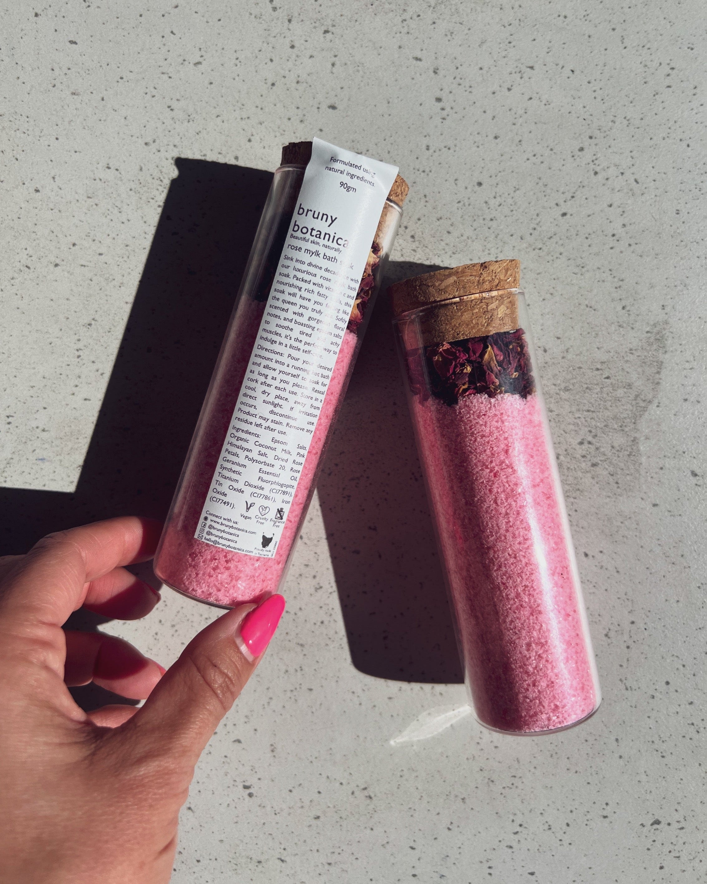 Hand holding a container of Rose Mylk Bath Soak on a grey surface