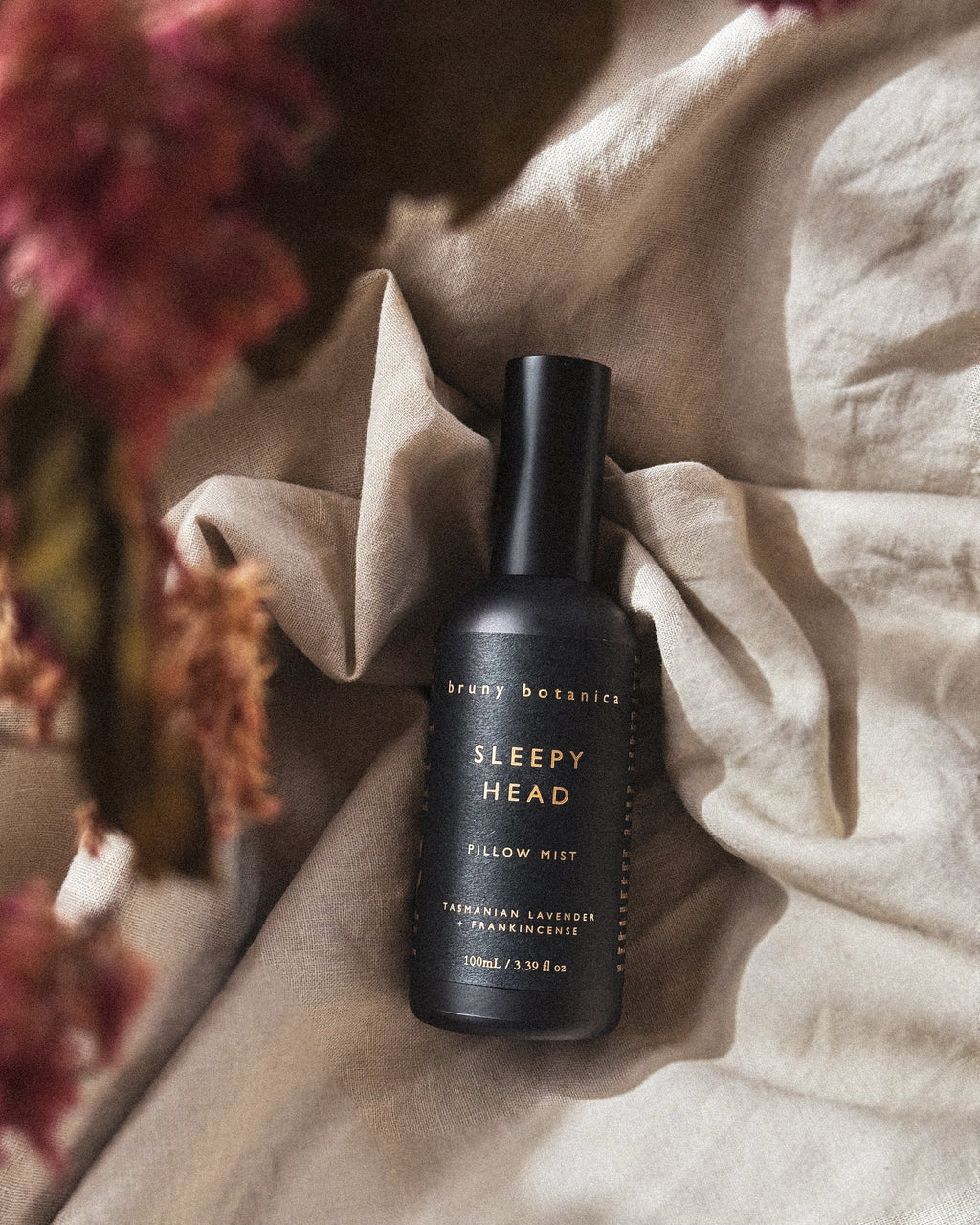 Black bottle of 'Sleepy Head' pillow mist on a textured surface with flowers in the background