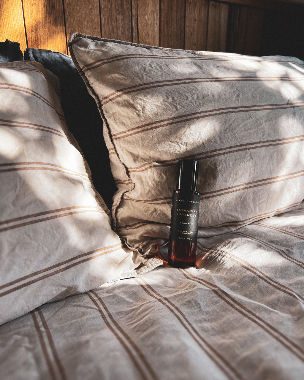 A Tasmanian Lavender Room Mist bottle on a bed with striped bedding and wooden headboard