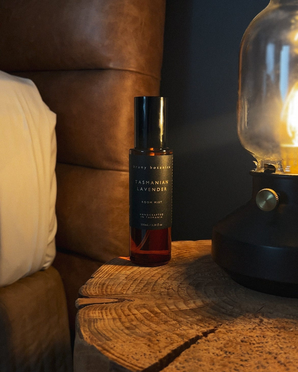 Small bottle of Tasmanian Lavender Room Mist on a wooden nightstand next to a lamp with a warm glow.