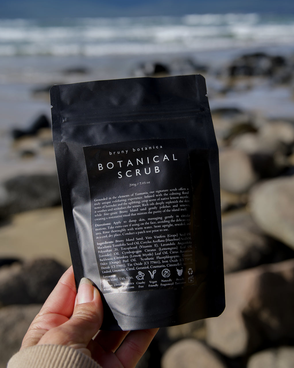 Hand holding a package of the Bruny Botanica Botanical Scrub with a beach background
