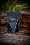 Black packaging of Bruny Botanica's Botanical Scrub on a wooden surface with blurred greenery in the background