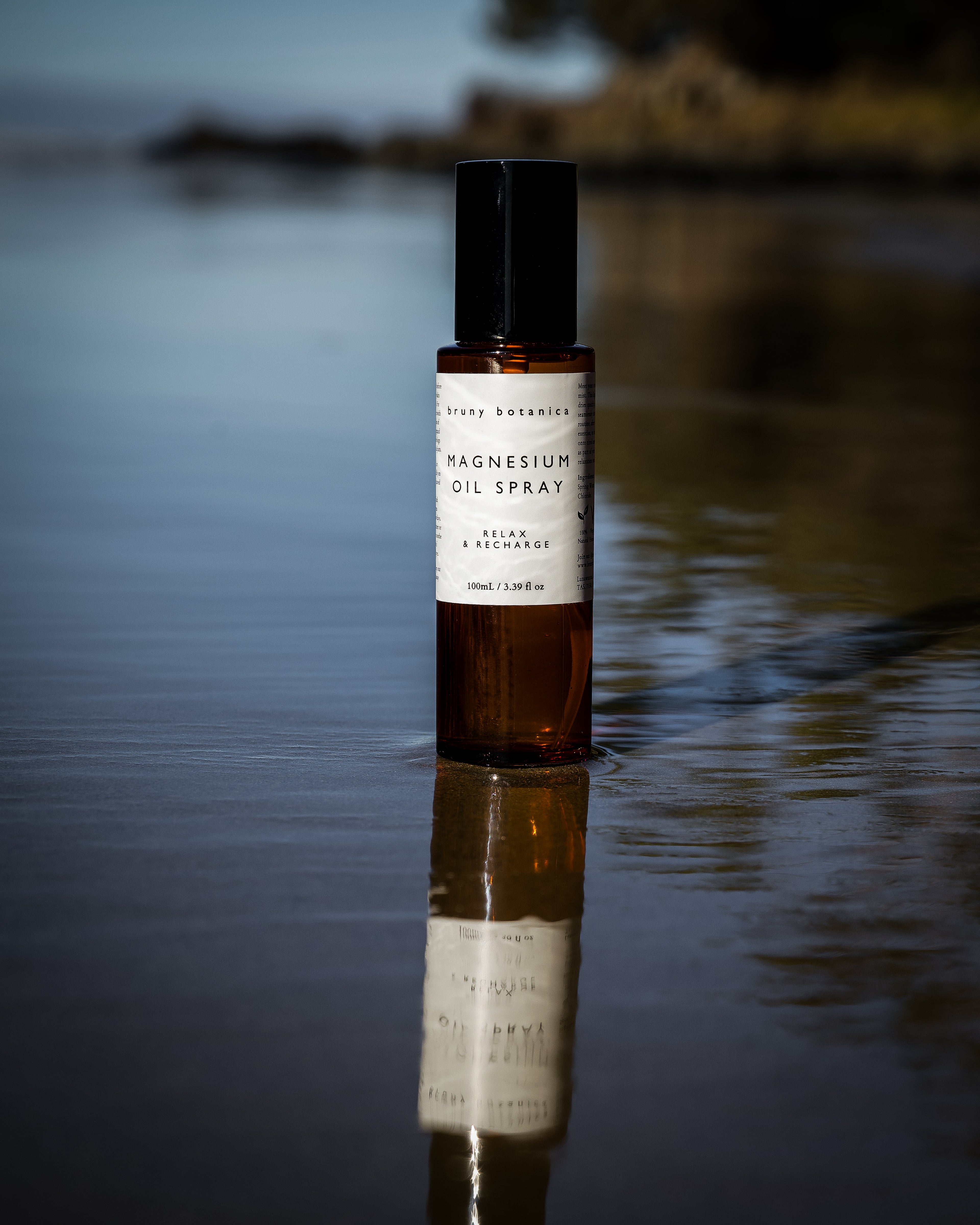 A bottle of Bruny Botanica's Magnesium Oil Spray stands in calm water, with its reflection visible on the surface. The amber glass bottle has a black cap and a white label with black text.