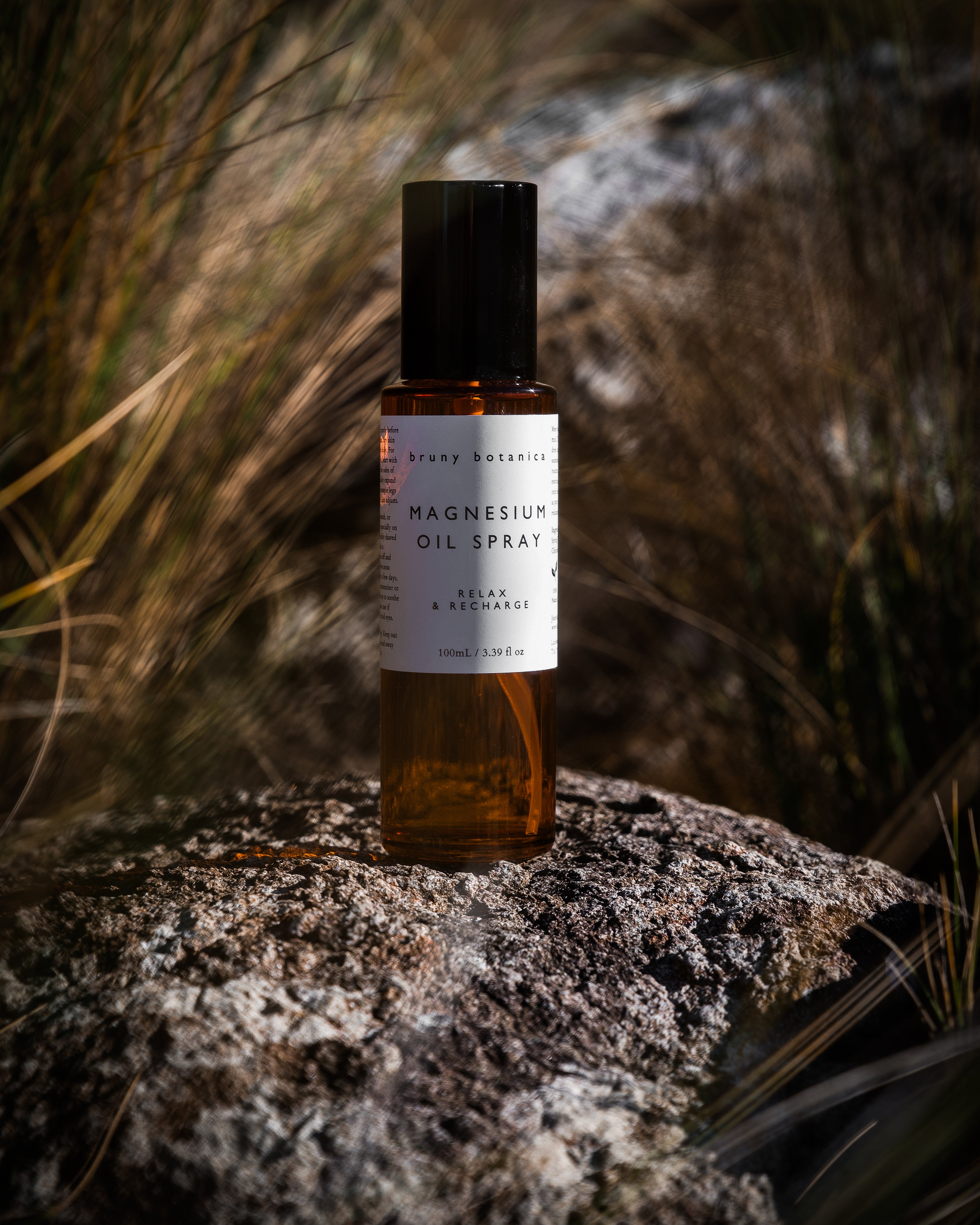 A bottle of Bruny Botanica's Magnesium Oil Spray stands on a textured rock outdoors, surrounded by dry grass and natural light.