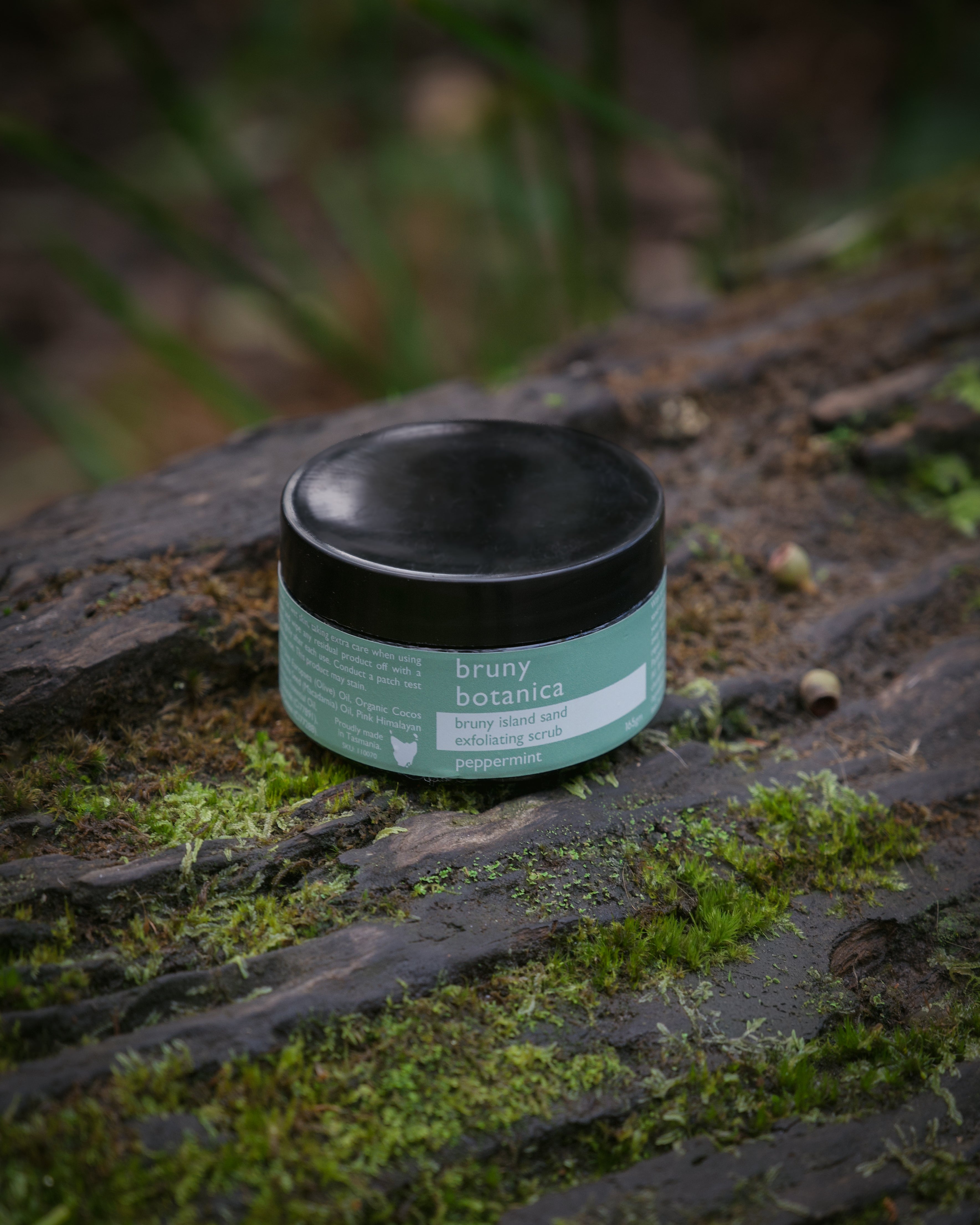A jar of Bruny Island Sand Exfoliating Scrub Peppermint sits atop a mossy log in the forest